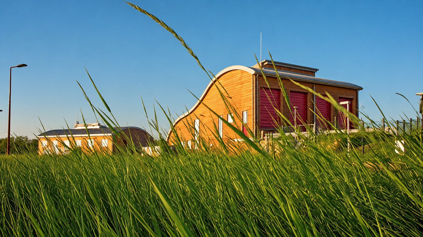 Un bâtiment moderne en bois au toit arrondi et aux volets rouges se dresse sous un ciel bleu, vu à travers de hautes herbes au premier plan.