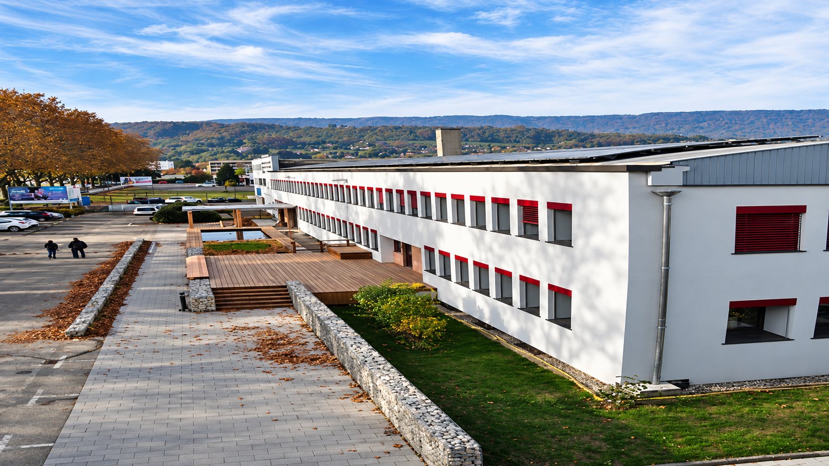 Vue d’un long bâtiment blanc à fenêtres encadrées de rouge, avec une terrasse en bois, un muret de pierre et une pelouse, au pied de collines.