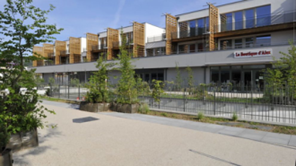 Un bâtiment contemporain à plusieurs étages avec des éléments de façade en bois, une enseigne rouge au rez-de-chaussée et une allée en gravier au premier plan.