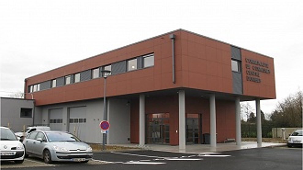 Un bâtiment moderne à deux niveaux, aux façades rouge brique et grise, avec des voitures garées devant et un panneau de texte sur le mur de droite.