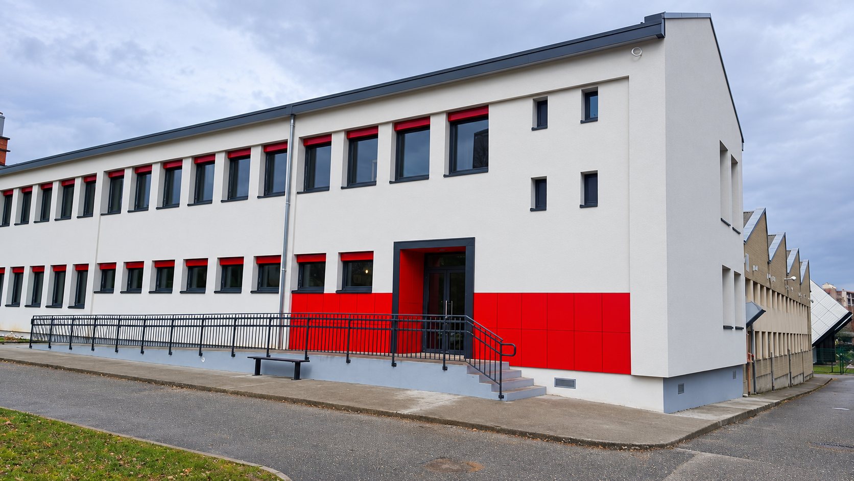Façade d’un bâtiment administratif récemment rénové, blanche avec des panneaux rouges autour de l’entrée, une rampe d’accès et un ciel nuageux.