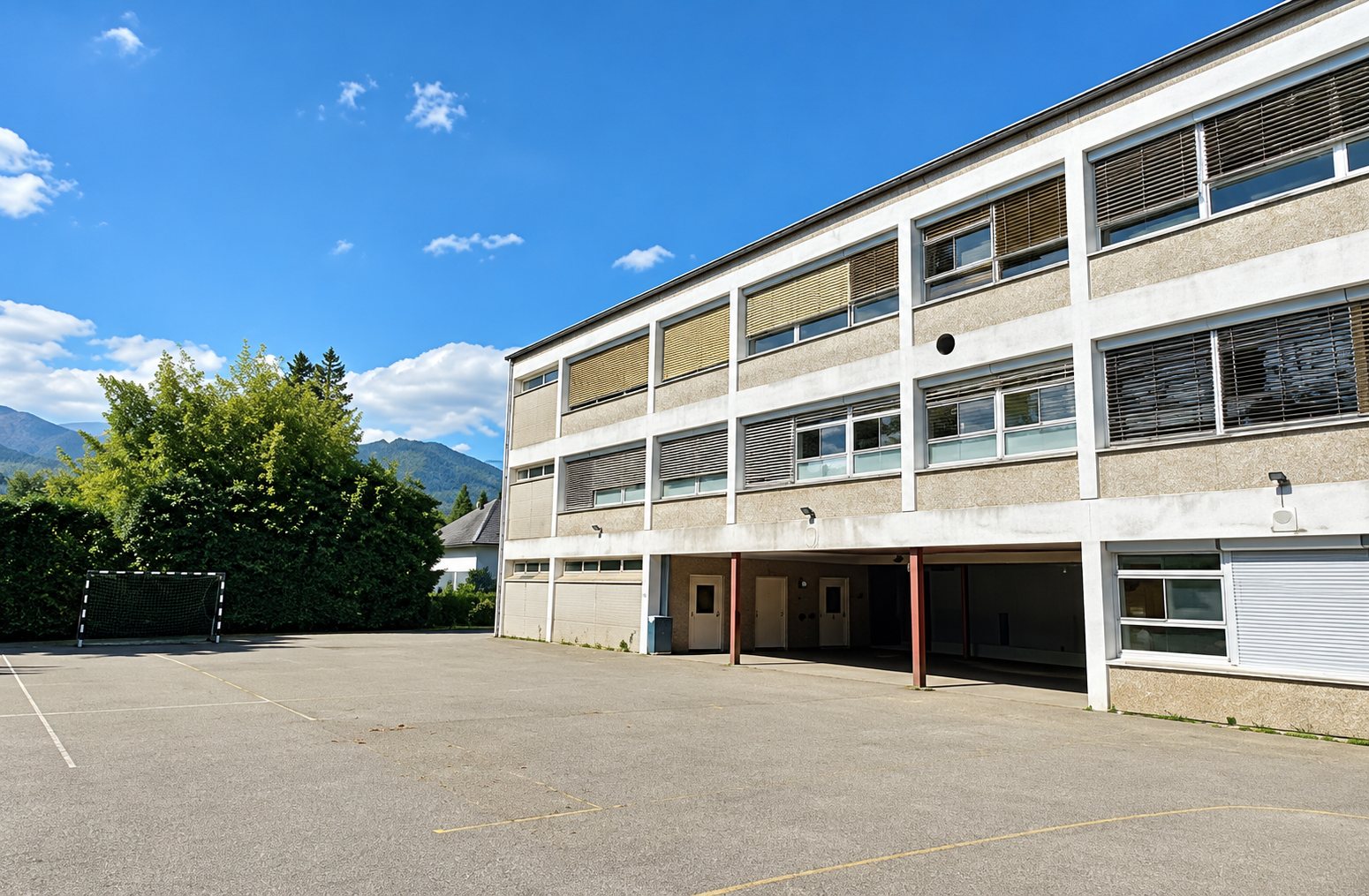 Un bâtiment à plusieurs étages aux façades beige donne sur une grande cour goudronnée, avec des arbres et des montagnes sous un ciel bleu.