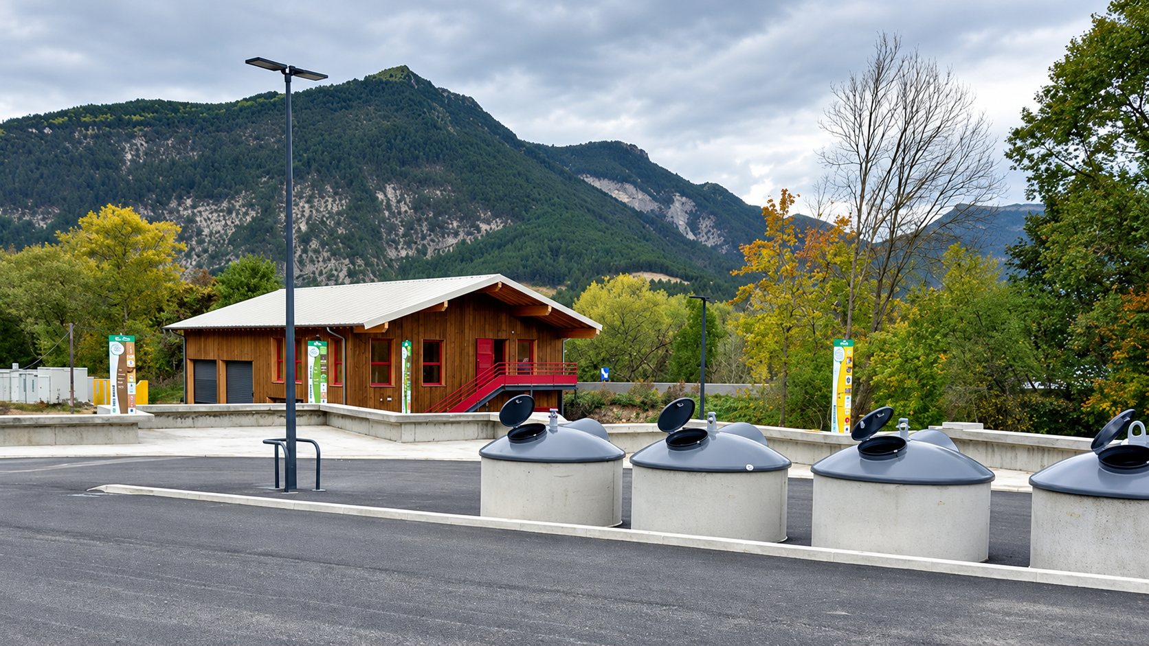 Quatre conteneurs à déchets semi-enterrés alignés sur un parking récemment goudronné, devant un chalet en bois et une montagne boisée.
