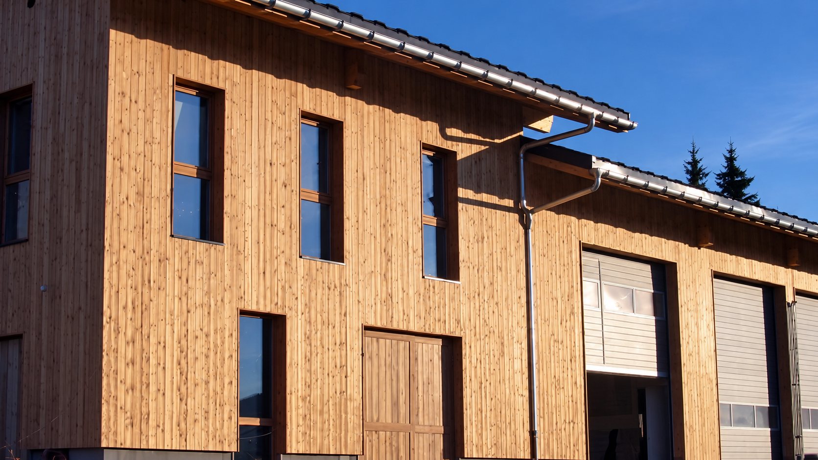 Façade d’un bâtiment moderne recouvert de bardage en bois vertical, avec fenêtres étroites, grandes portes coulissantes et toit en pente, éclairée par le soleil.