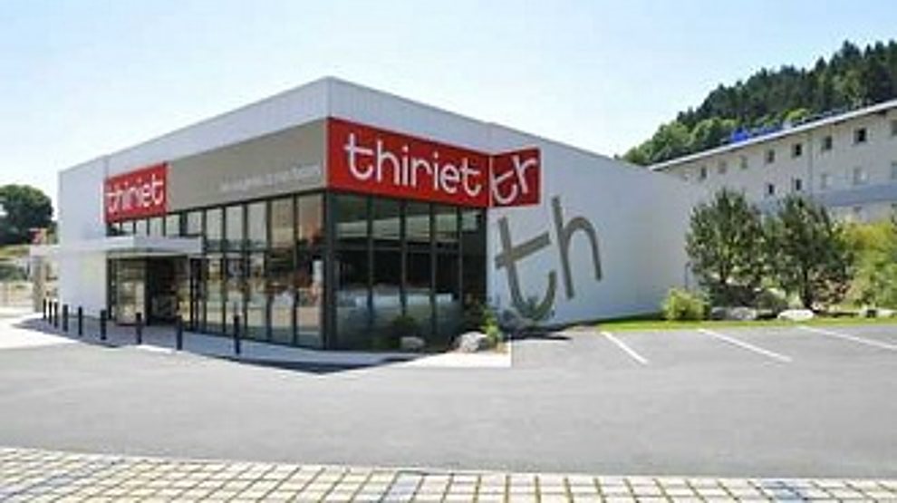 Façade vitrée d’un magasin Thiriet avec son enseigne rouge et blanche, un parking devant et des collines boisées en arrière-plan.