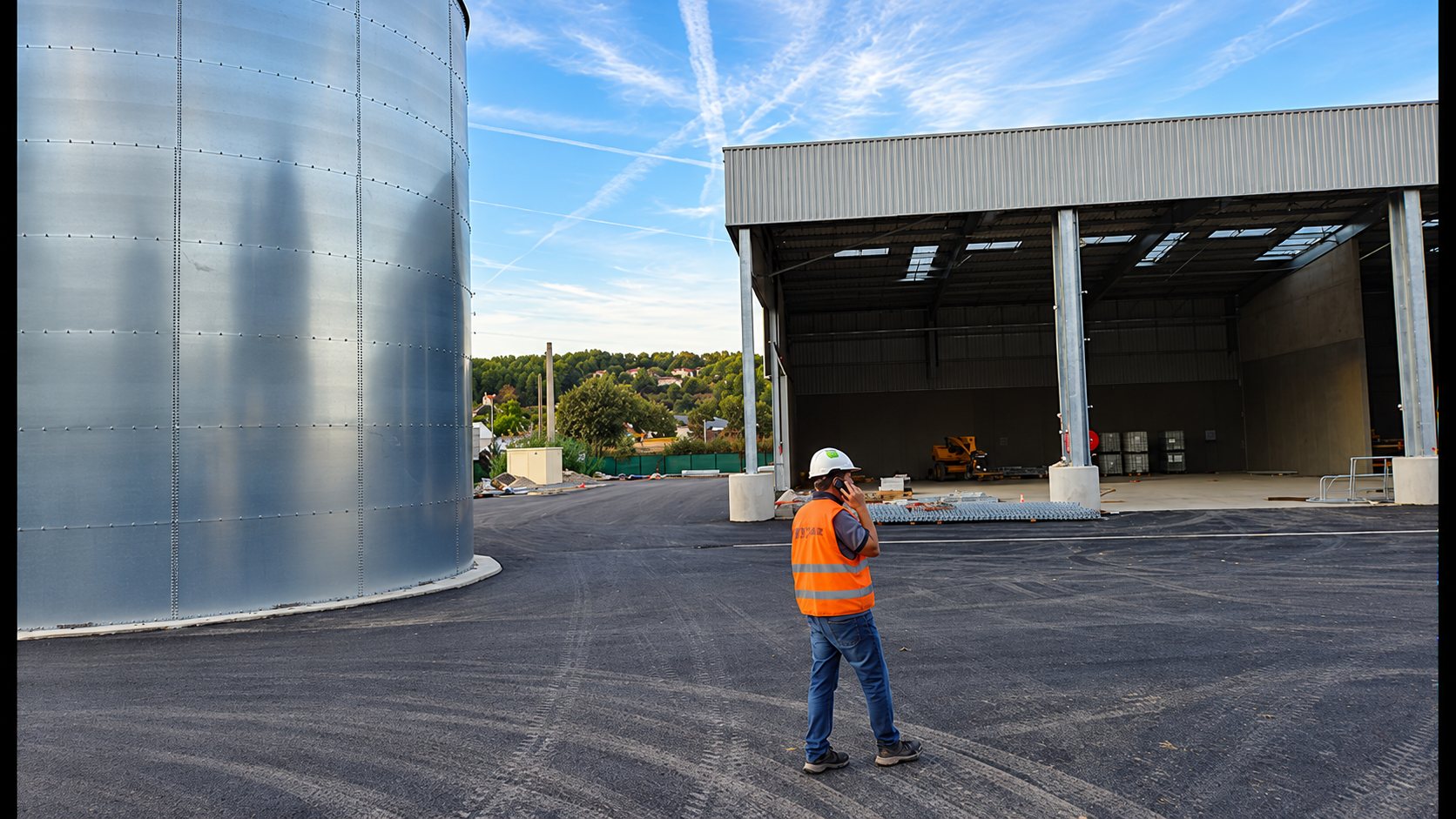 Un ouvrier casqué en gilet orange téléphone sur un chantier, entre un grand réservoir en métal galvanisé et un hangar industriel en construction.