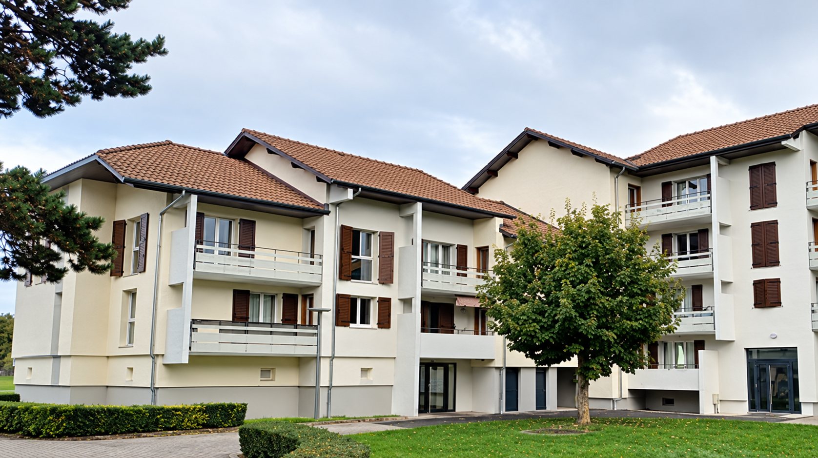 Un immeuble résidentiel de deux ou trois étages aux façades beige clair, avec balcons et volets bruns, entouré de pelouse et d’arbres sous un ciel nuageux.